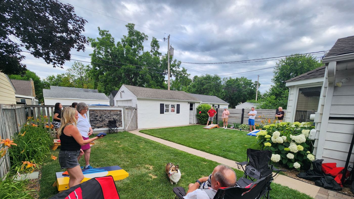 A backyard cornhole night: two boards on the grass, players mid-game, a corgi watching.